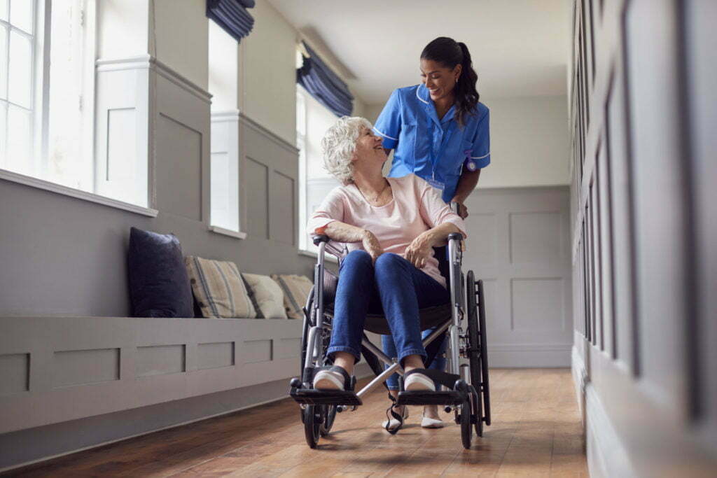 Senior Woman At Home Being Pushed In Wheelchair By Female Care Worker In Uniform